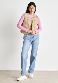 Beige bomullsvest med trykknapper, brukt over en lys rosa ribbestrikket langermet skjorte. Kombinert med lyseblå rette jeans og hvite joggesko.