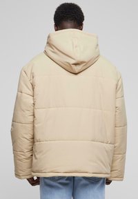 Beige puffjacka med huva, som har en quiltad design och lös passform. Tyget verkar mjukt med en slät textur.