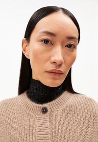 Beige gebreide cardigan met een ronde halslijn en een enkele zwarte knoop, gedragen over een zwarte geribbelde coltrui. Gladde textuur.