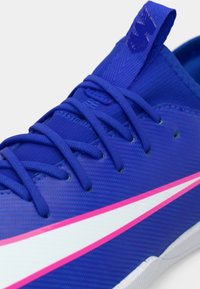 Modrá sportovní bota s fialovými tkaničkami a bílým svíčkovým logem obrysem v růžové na boku, s texturovaným textilním svrškem.