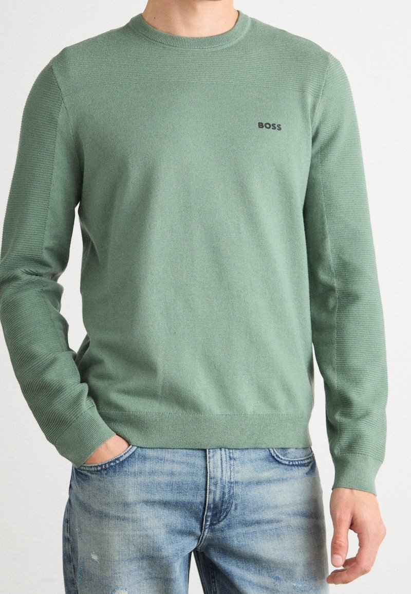 Homme portant un pull ras-du-cou vert clair avec des manches côtelées et un logo noir "BOSS", associé à un jean délavé bleu clair.