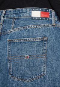 Zili zili džinsi ar aizmugurējo kabatu, kurā ir neliels Tommy Jeans logotipa uzlīme. Jostasvieta ir apzīmogota ar zīmola etiķeti sarkanas, baltas un zilas krāsās.