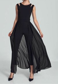 Frau in einem ärmellosen schwarzen Jumpsuit mit transparenter, fließender Überrock, die mit gespreizten Füßen steht und die Kanten des Rocks hält.