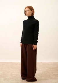 Kobieta z czerwonymi włosami, ubrana w czarny sweter typu golf i szerokie brązowe spodnie, stoi na beżowej podłodze przed gładką ścianą.