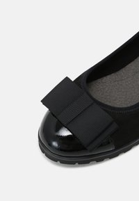 Chaussure plate noire avec embout verni et grand nœud en tissu texturé, intérieur rembourré et semelle en caoutchouc durable.