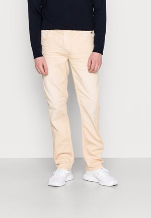 Homme portant un pantalon beige, des baskets blanches et une chemise noire à manches longues, debout devant un fond clair uni.