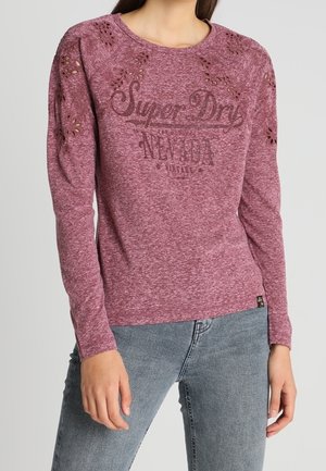 Vrouw die een paarse gebreide top met lange mouwen draagt, met bloemvormige uitsparingen en de tekst "Super Dry Nevada", gecombineerd met lichtblauwe spijkerbroek.