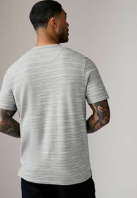 Cămașă gri cu mânecă scurtă, cu un model texturat în dungi, gât rotund și o croială lejeră. Prezintă o margine dreaptă și fără branding vizibil.