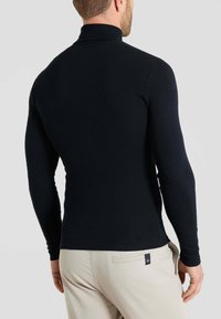 Maglietta nera a maniche lunghe con collo alto, dal design aderente, realizzata in tessuto morbido, abbinata a pantaloni di colore chiaro. Nessun motivo o accento visibile.