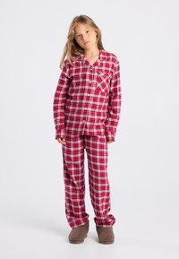 Meisje in rood en wit geruite pyjama en bruine pantoffels tegen een effen grijze achtergrond.
