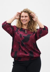 Blouse noire avec des motifs floraux rouges, encolure ronde avec un détail en clé, et manches trois-quarts. Fabriquée en tissu lisse.
