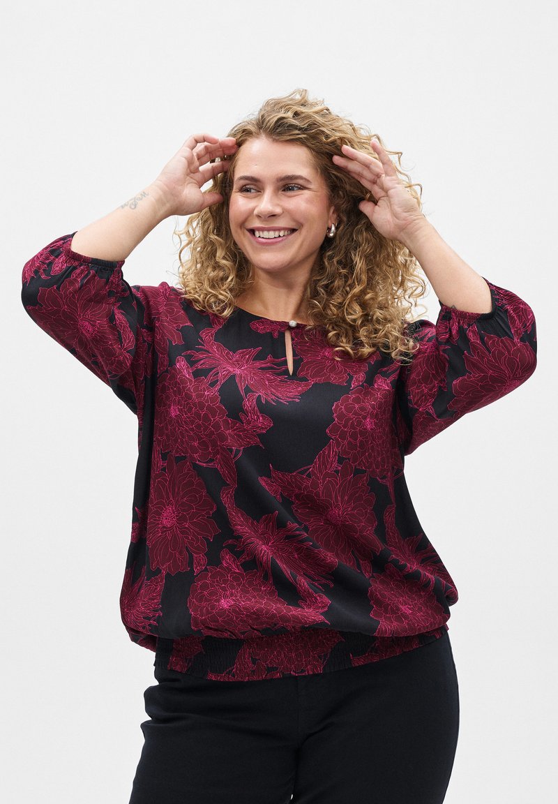 Blouse noire avec des motifs floraux rouges, encolure ronde avec un détail en clé, et manches trois-quarts. Fabriquée en tissu lisse.