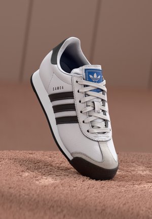 Hvid Adidas Samoa sneaker med sorte striber, blåt logo på tungen og sort sål på en brun tekstureret overflade.