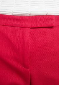 Milieu du corps d'une personne portant un pantalon rouge avec une chemise blanche texturée rentrée, montrant la ceinture et la zone de la braguette.