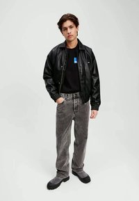 Jachetă bomber din piele ecologică neagră purtată peste un tricou negru, asociată cu blugi gri din denim și botine groase negre. Design simplu, croi lejer.