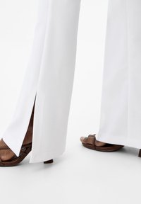 Vita vida byxor med sidoklyftor, i kombination med bruna öppna klacksandaler. Den släta tyget och den skräddarsydda designen framhäver silhuetten.