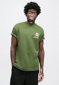 Maslinasto zelena pamučna majica kratkih rukava s podvrnutim rukavima. Na prsima se nalazi mali bijeli grafički prikaz s tekstom i kružnim dizajnom.