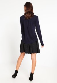 Femme aux cheveux bruns et ondulés portant un chemisier bleu marine à manches longues, une jupe texturée noire et des bottines noires, debout de dos.