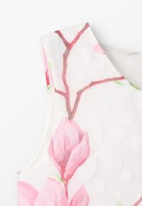 Tílko bez rukávů s květinovým vzorem na bílém podkladu, zdobené růžovými květy magnólie a zelenými akcenty, vyrobené z lehké texturované látky.