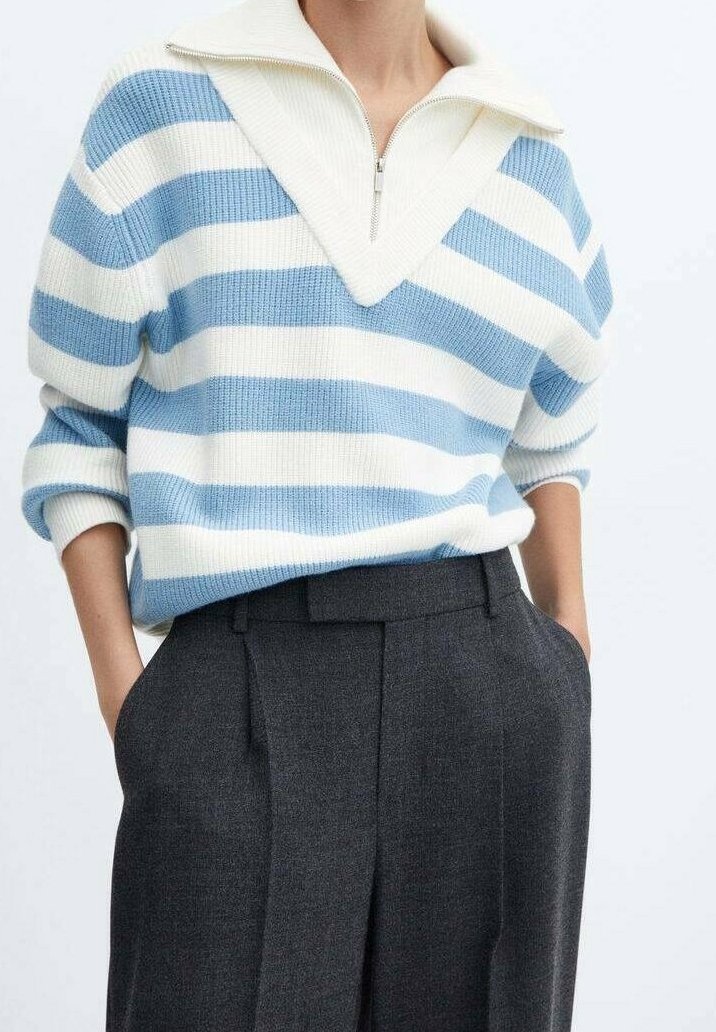 Personne portant un pull en maille rayé blanc et bleu clair avec un col zippé et un pantalon taille haute gris foncé, mains dans les poches.