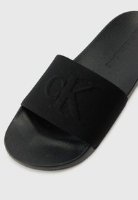 Black slide sandal featuring a matte fabric strap embossed with "CK" and a textured rubber sole, offering a minimalistic design.