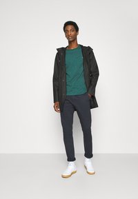 Sort vandafvisende jakke over en grønmønstret langærmet skjorte, marineblå bukser og hvide sneakers med gummisåler, stående foran en ensfarvet baggrund.
