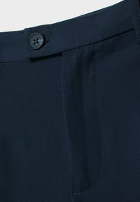 Gros plan sur la ceinture d'un pantalon bleu marine foncé avec une fermeture à bouton et des coutures visibles sur la poche latérale.