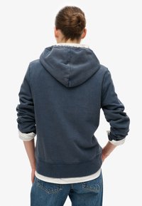 Sudadera azul marino con bolsillo tipo canguro, puños y dobladillo acanalados. Presenta una textura suave y una capucha forrada. Usada sobre una camiseta blanca.