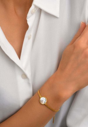 Femme portant un bracelet en or avec un centre rond en nacre à son poignet, vêtue d'une chemise blanche boutonnée.