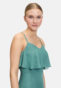 Robe en sarcelle avec de fines bretelles et un vol à plusieurs couches au niveau de la poitrine. Texture lisse semblable à du satin, avec un corsage ajusté et une partie inférieure évasée.