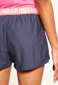 Námořnické sportovní šortky s elastickým růžovým pasem, na kterém je vytištěno "UNDER ARMOUR" bílou barvou. Hladká textura, funkční design a boční rozparky.