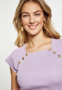 Top ribbed lavandă cu mâneci scurte, având butoane decorative aurii pe de-a lungul gâtului. Textură fină și design mulat.
