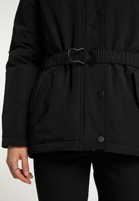 Chaqueta acolchada negra con cintura ceñida, dos bolsillos laterales y cierres de botón a presión. Tela lisa con acabado mate.