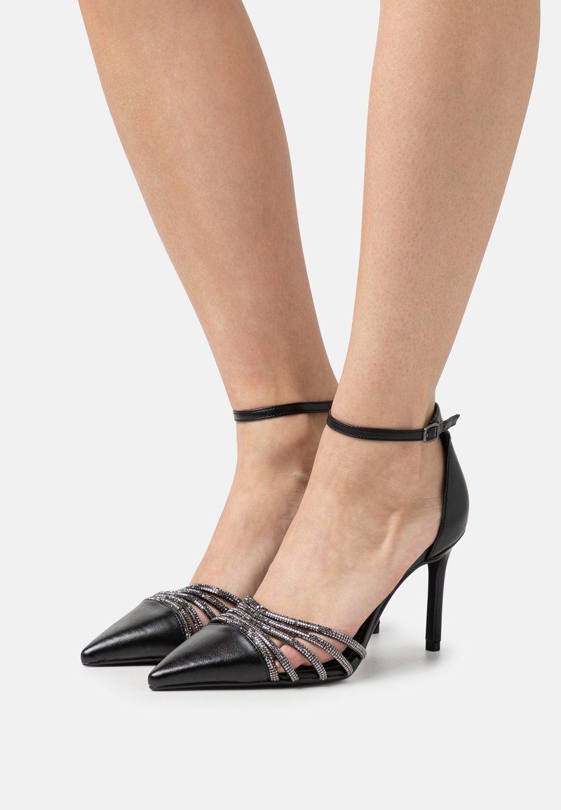 Chaussures noires à talons hauts avec bout pointu, bride autour de la cheville et lanières métalliques décoratives traversant le dessus du pied pour plus de détails.