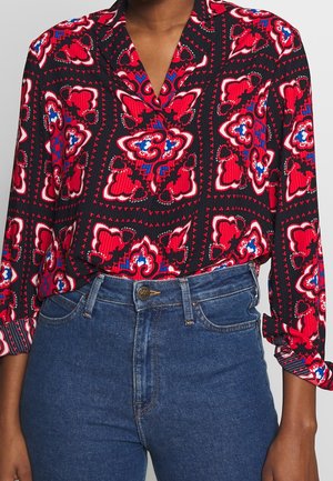 Blouse à motifs rouges et bleus sur fond noir, avec un col et des manches longues. Associée à un jean bleu taille haute.