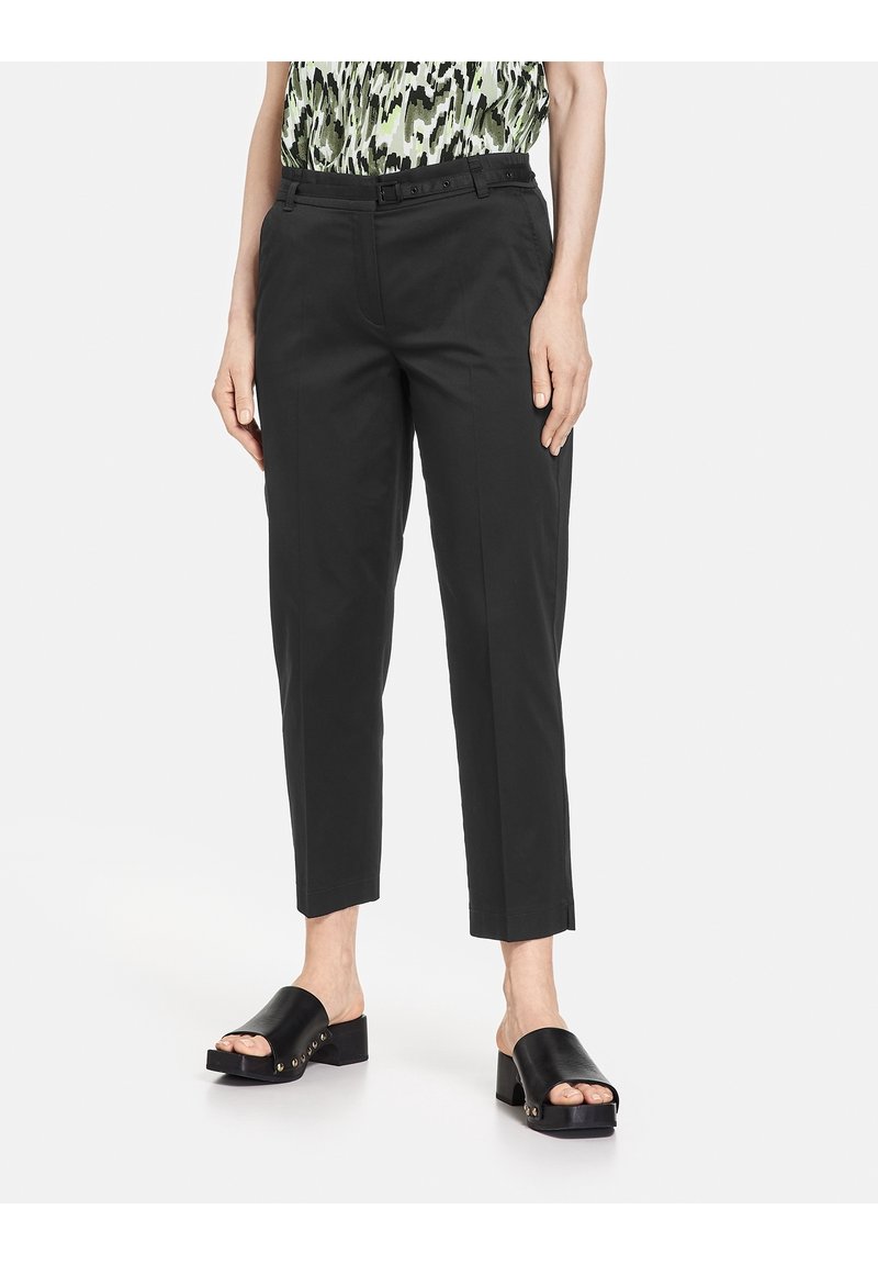 Femme portant un pantalon noir coupé sur mesure avec boucle latérale réglable et des sandales noires à talons blocs ouvertes.
