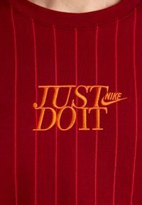 Sarkans īsām piedurknēm krekls ar vertikālām svītrām, kuram ir izšūts dzeltenais un oranžais "JUST DO IT" logo un Nike svītra.