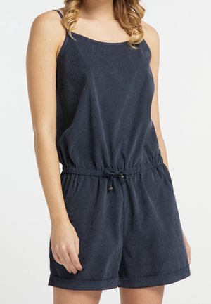 Femme portant une combinaison-short sans manches bleu foncé avec une taille à cordon ajustable et des poches, debout devant un fond uni.