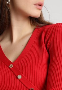 Czerwony prążkowany sweter z dzianiny z dekoltem w szpic, zdobiony guzikami z srebrnymi akcentami, o dopasowanym kroju podkreślającym fakturę i kształt.