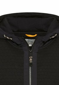 Veste à capuche noire avec un motif texturé en nid d'abeille, dotée d'une touche d'orange, d'une fermeture éclair et de cordons de serrage ajustables sur la capuche.