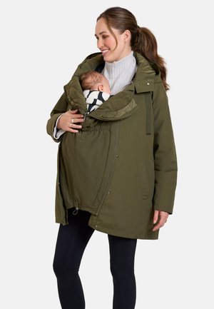 Vrouw die een olijfgroene draagjas draagt en een slapende baby houdt, gewikkeld in de jas.