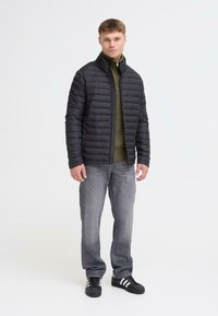 Sort pufferjakke med vandret syninger, høj krave og lynlås foran. Båret over en grøn striktrøje og grå jeans. Sorte sneakers.