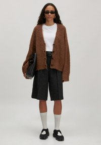 Bruine fluwelen cardigan, witte t-shirt, op maat gemaakte zwarte shorts, witte sokken, zwarte schoenen; houdt een zwarte handtas vast; relaxte uitstraling.