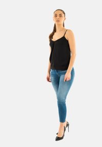 Camisole noir avec des accents cloutés, associé à un jean ajusté bleu clair et des talons aiguilles noirs, se tenant sur un fond uni.