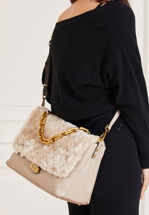 Beigetöne Lederhandtasche mit einer flauschigen beigen Pelzklappe, ausgestattet mit einem goldfarbenen Kettenriemen und einem dezenten Verschlussdetail, über der Schulter getragen.