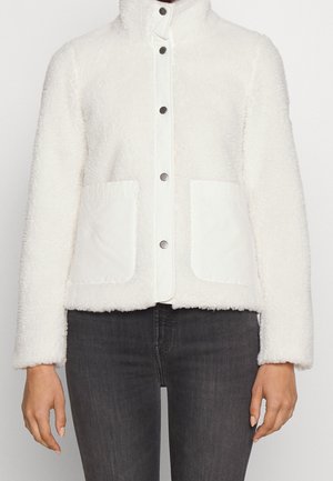 Witte fleece jas met knopen en twee voorzakken, gedragen met donkergrijze spijkerbroek, zichtbaar van nek tot heupen.