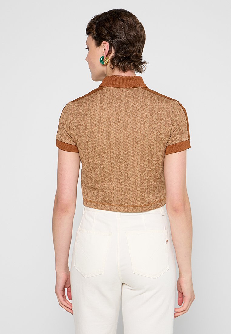 Polo de color marrón con estampado, de manga corta, diseño texturizado en diamante y cuello sólido. Llevado con pantalones blancos de cintura alta.