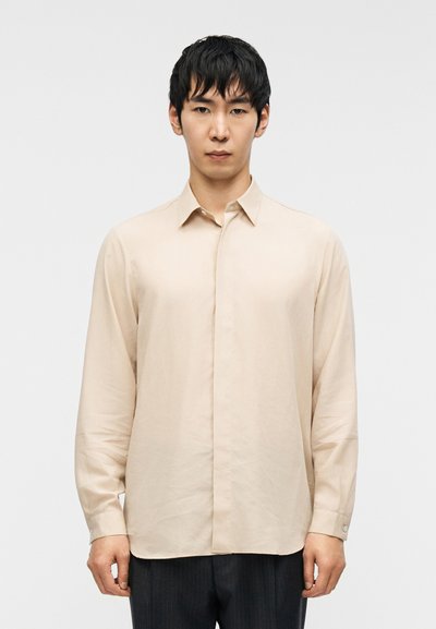 Chemise manches longues beige clair, en tissu doux, avec une fermeture boutonnée sur le devant, un col pointu et des poignets à un bouton. Design minimaliste.