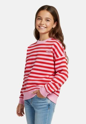 Glimlachend meisje met lang bruin haar draagt een roze-rood gestreepte trui en lichtblauwe jeans, hand in de zak, poserend tegen een witte achtergrond.