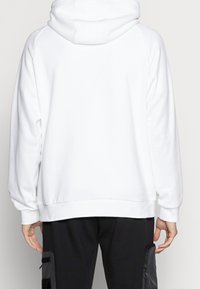 Vit hoodie med en mjuk textur, huva och raglanärmar. Har en avslappnad passform och ribbad nederkant. Bärs över svarta byxor med fickor.